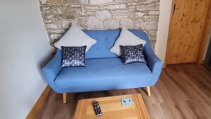 Een blauwe bank met gekleurde kussens, een afstandsbediening en een onderzetter op een houten salontafel in een kamer met houten vloeren en een stenen muur.