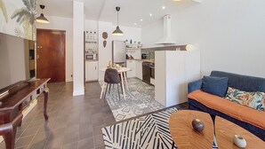 Un salon moderne avec un canapé, une table basse et un piano. Au fond, on aperçoit une cuisine équipée d’un réfrigérateur, d’un four à micro-ondes et de placards.