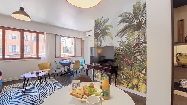 Un salon moderne comprenant un coin repas, de grandes fenêtres, un téléviseur à écran plat et une fresque murale aux motifs tropicaux.
