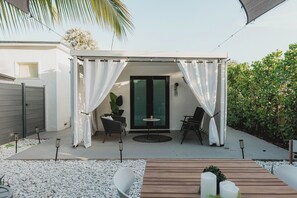 A covered outdoor seating area with two chairs and a table, surrounded by white pebbles and greenery.