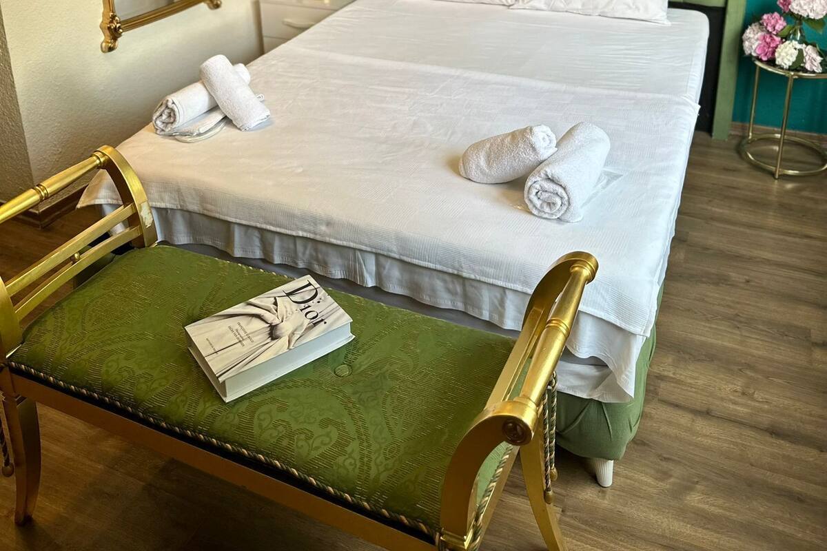 Habitación de hotel con una cama, un sillón tapizado en verde, un espejo con marco dorado y una mesita de noche con lámpara.