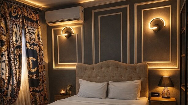 Un dormitorio con cabecera capitonada, una cama grande con ropa de cama blanca, una mesita de noche con lámpara y apliques decorativos en la pared.