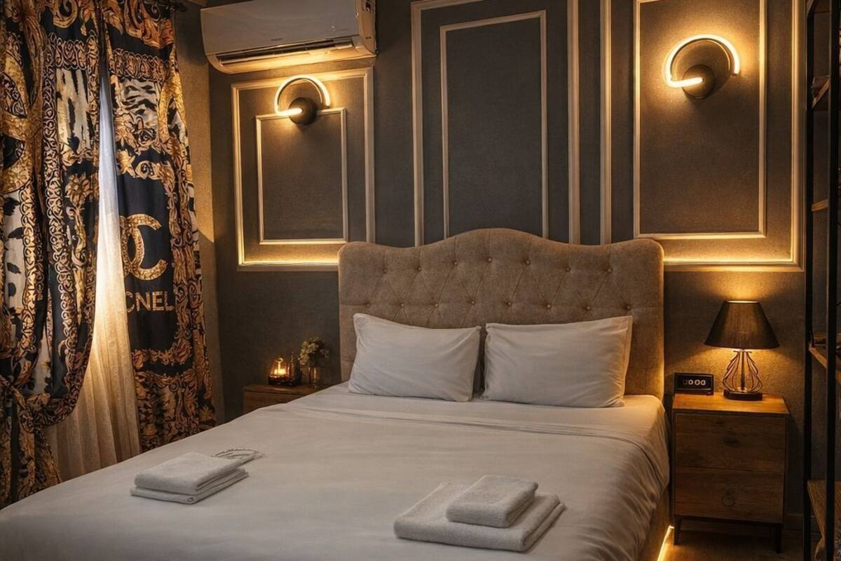 Un dormitorio con cabecera capitonada, una cama grande con ropa de cama blanca, una mesita de noche con lámpara y apliques decorativos en la pared.