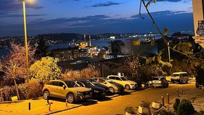 Un área de estacionamiento con varios coches, una farola y vistas de un paisaje urbano con edificios y una masa de agua al anochecer.