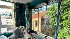 Habitación de hotel con una cama, una silla, una mesita con un florero y vistas a edificios y árboles a través de ventanas amplias.