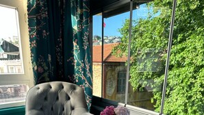 Uma cama bem arrumada com uma toalha branca, uma poltrona cinza estofada, uma pequena mesa redonda com um vaso de flores e vista para um prédio de telhado vermelho através de uma janela.