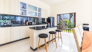 A modern kitchen with a black countertop, wooden cabinets, and a glass backsplash. There are two bar stools and a dining area with a view of the outdoors. - Villa Utara - Two Bedroom Villa, Sleeps 4 (Seminyak)