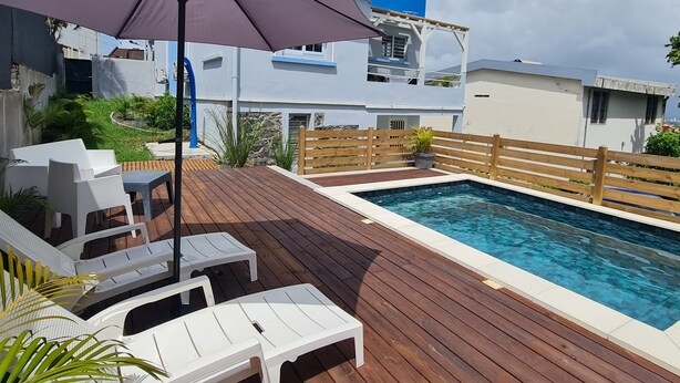 A swimming pool area with a wooden deck, white lounge chairs, and a blue building in the background. - VBF -studio Pool View (Fort-de-France Bay)