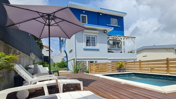 A blue and white two-story house with a wooden deck, outdoor furniture, and a swimming pool. - VBF - Cosy Apartment (Fort-de-France Bay)