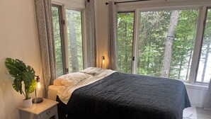 A bedroom with a bed, a bedside table with a lamp, a potted plant, and large windows with a view of trees. - New 2-bedroom cottage in Bancroft Ontario, fully renovated on the lake (MONT)