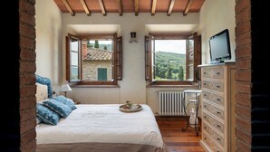 A bedroom with a bed, dresser, TV, and two windows with views of a stone building and greenery.