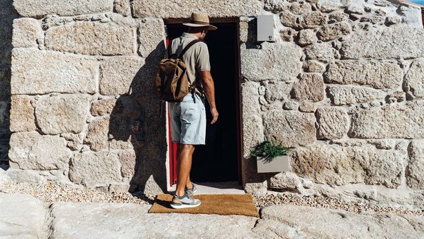 A man standing at the entrance of a stone building with a backpack, a hat, and shorts. - a small, very elegant and cosy studio loft of almost 14m2 (Sortelha)