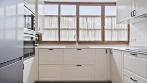 A modern kitchen with white cabinetry, a built-in oven, a window with curtains, and a stainless steel refrigerator.