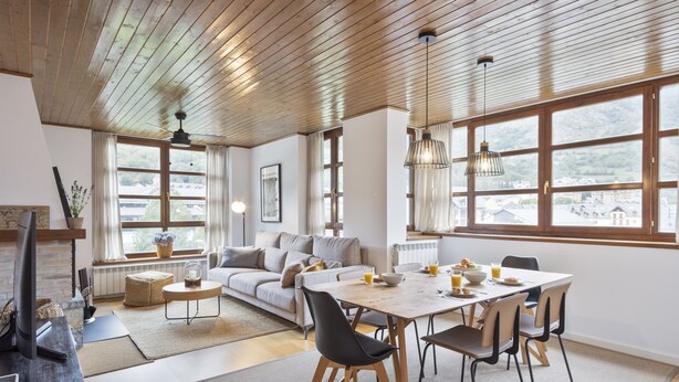 A modern living room with a wooden ceiling, large windows, a dining area with a table and chairs, and a sofa.