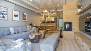 Un salon moderne avec un canapé, une table basse et un coin repas avec des chaises. On aperçoit une cuisine à l’arrière, avec une cuisinière et une table à manger.