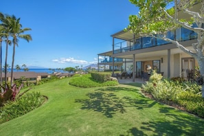 A modern two-story house with a covered patio, lush green lawn, and a view of the ocean.