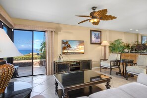 A living room with a large window, a ceiling fan, a fireplace, and a dining area. - Wailea Elua Village | Ocean View 2 BR | Car Incl. w/ 6+ Nights | WEV-2009 by KBM (Wailea)