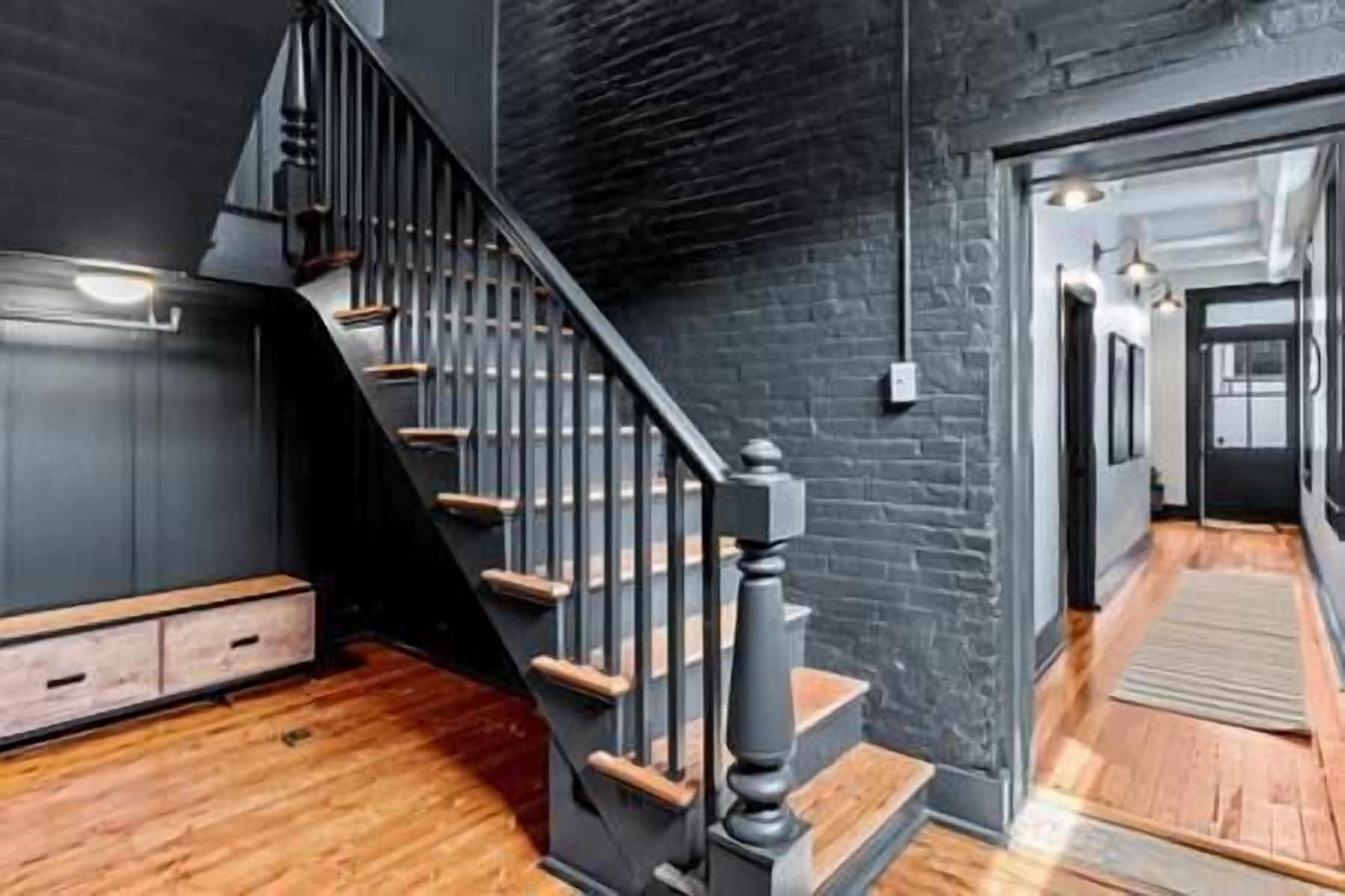 Un escalier avec des marches en bois et des rampes noires, menant à une pièce dont un mur est peint dans une couleur sombre et qui est éclairée par un luminaire suspendu.