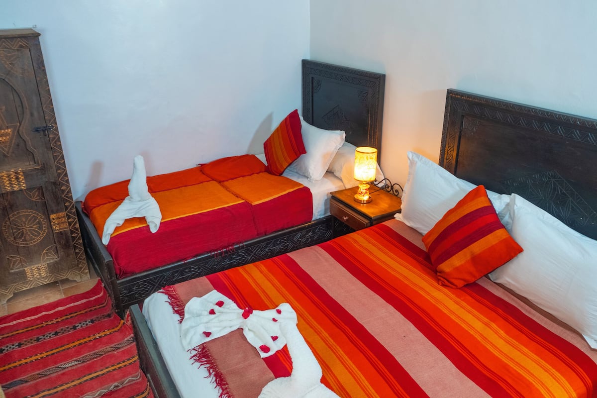 A room with two beds, each with red and orange bedding, a nightstand with a lamp, and a wooden cabinet with intricate carvings.