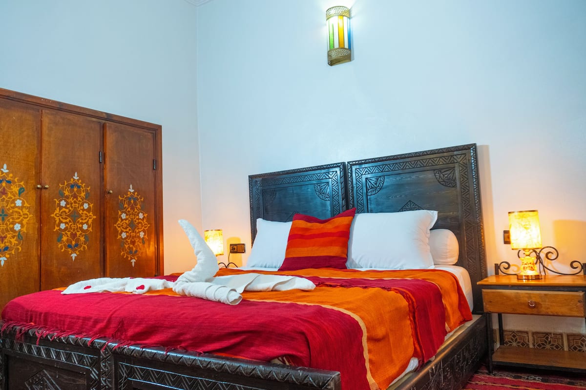 A hotel room with a large wooden wardrobe, a bed with a red and orange bedspread, white pillows, and two bedside lamps.