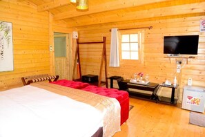 Habitación tipo cabaña de madera con cama, dos sillas rojas, un escritorio con computadora, televisor y un refrigerador pequeño.
