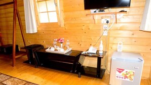 A wooden cabin interior with a wooden floor, a dining table with a tray of items, a small refrigerator, a television mounted on the wall, and a window with curtains.