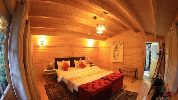 A wooden cabin bedroom with a bed, nightstands, a red ottoman, and a wooden ceiling.