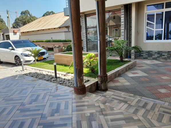 A building with a tiled entrance, a parked white car, and a small garden area with plants and a wooden planter.