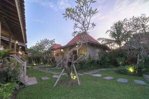 A well-maintained garden with stone pathways, a wooden staircase, and a building with a red roof. - Kirani Joglo (Celuk)