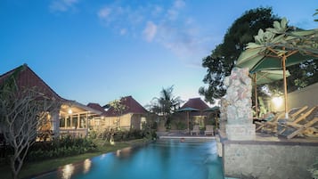 Zona de piscina con sillas de descanso y sombrillas, rodeada de edificios con techos rojos y cielo despejado.