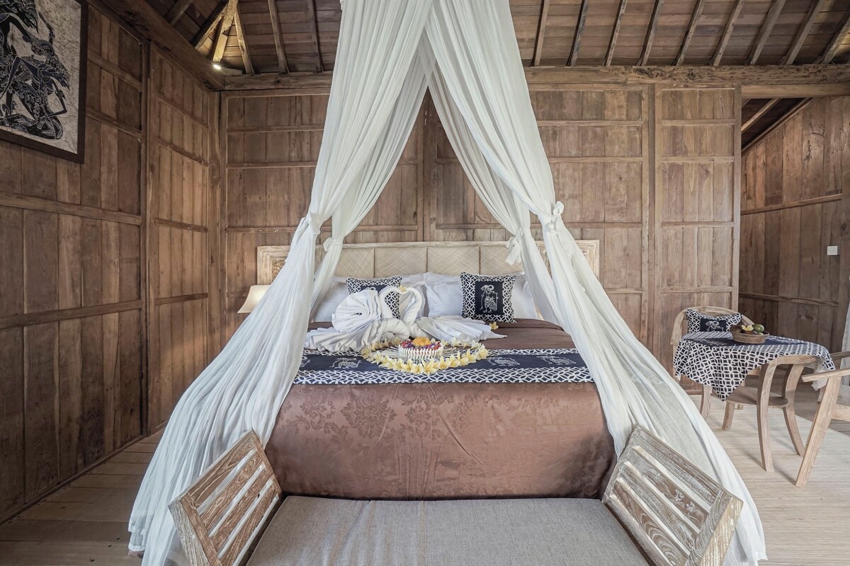 Una habitación de madera con una cama bajo un dosel, una mesa con mantel estampado y una silla.