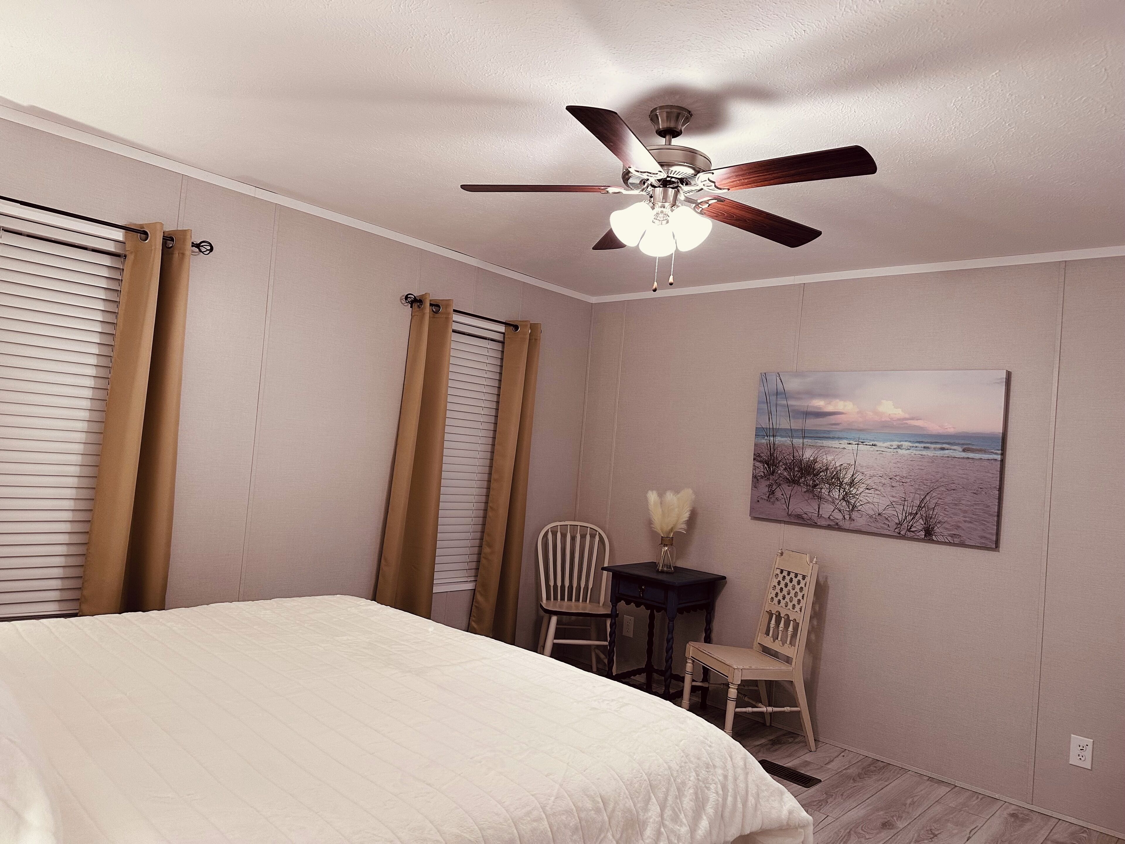 A bedroom with a bed, a ceiling fan, a window with blinds, a chair, a small table, and a wall-mounted painting.