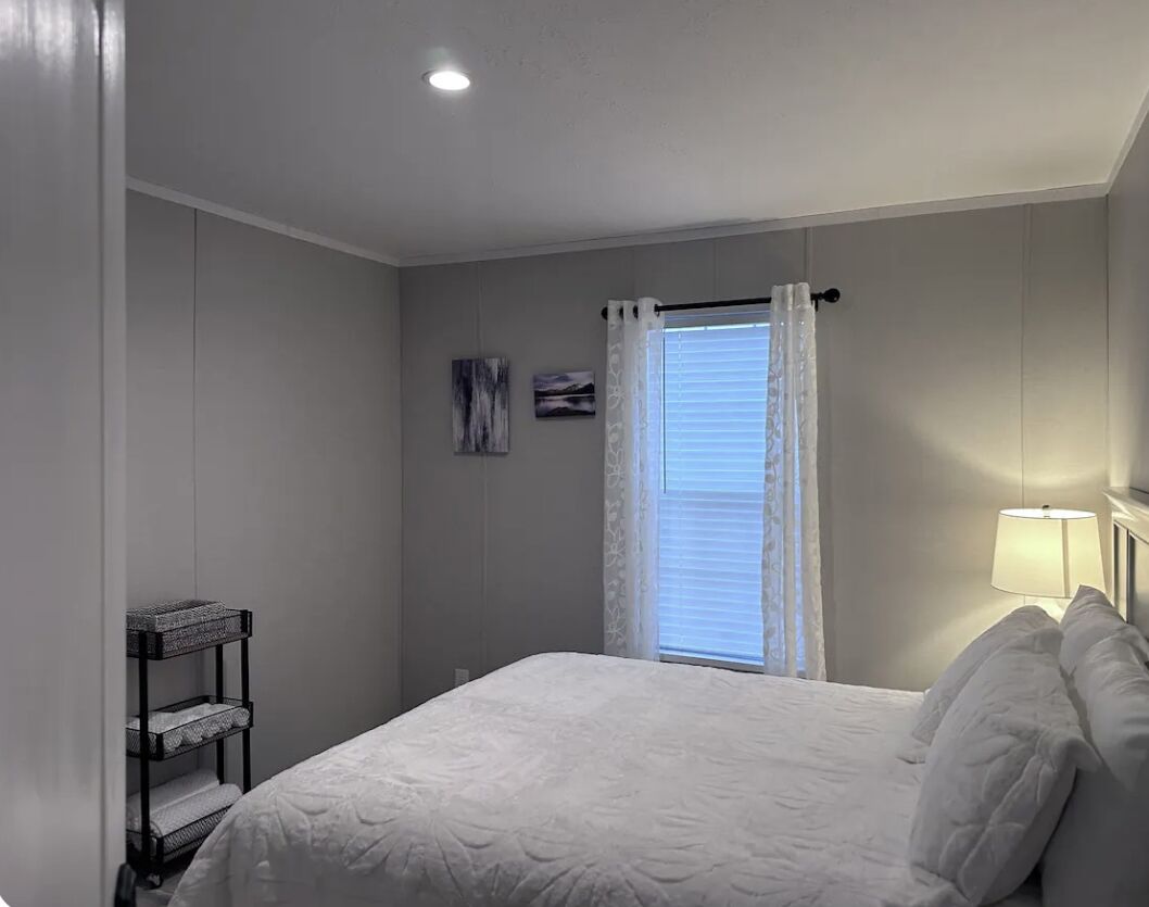 Un dormitorio con cama, mesita de noche, lámpara, ventana con cortinas y estante en la pared.