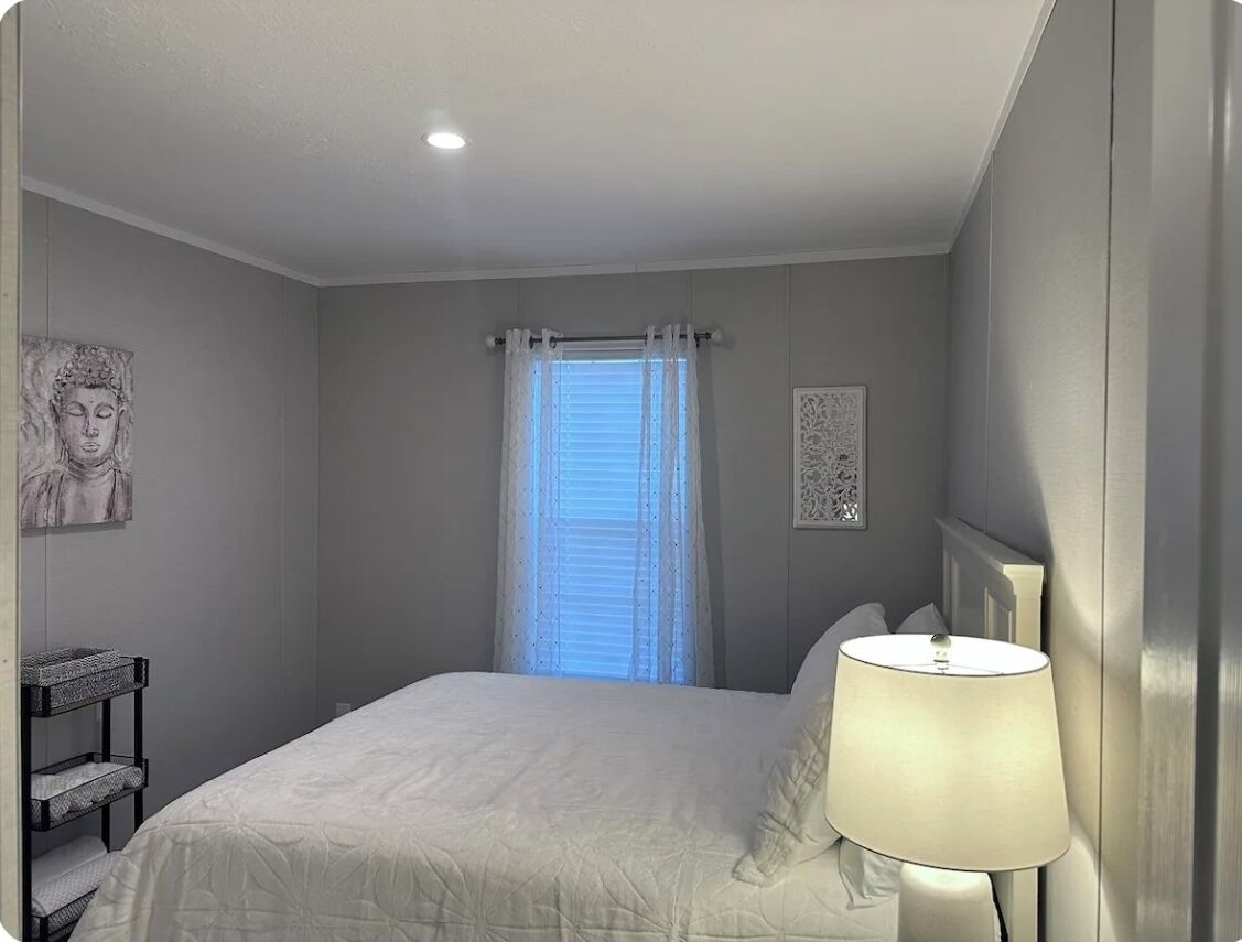 A bedroom with a bed, a bedside lamp, a window with curtains, and artwork on the walls.