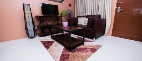 A living room with a sofa, a coffee table, a TV, a plant, and a window with curtains.