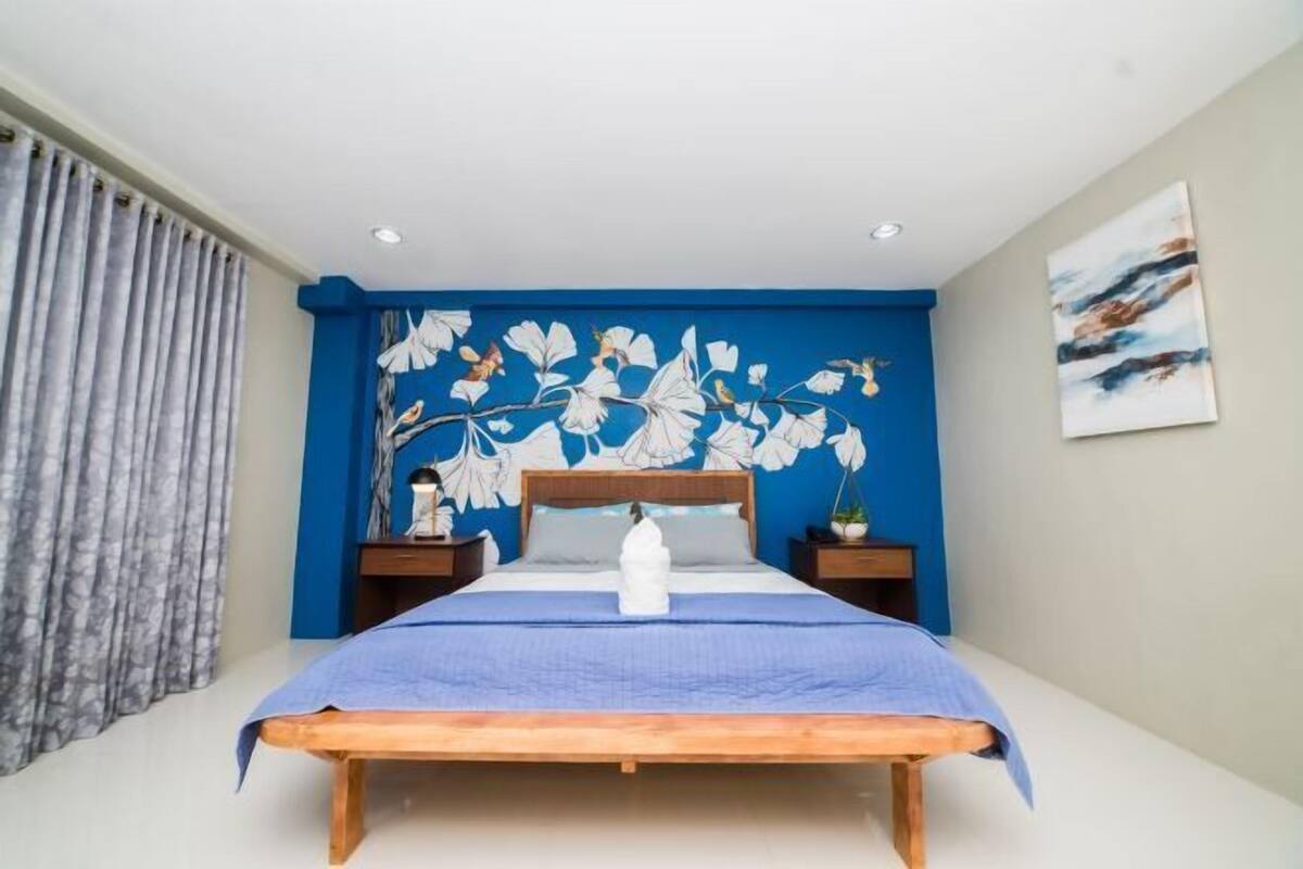 Un dormitorio con una cama, pared azul con detalles florales, un florero blanco y un cuadro en la pared.