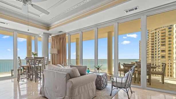 A spacious living room with a large sofa, a dining table, and chairs. The room has large windows offering a view of the ocean and a high-rise building. - La Playa 703 (Pensacola)
