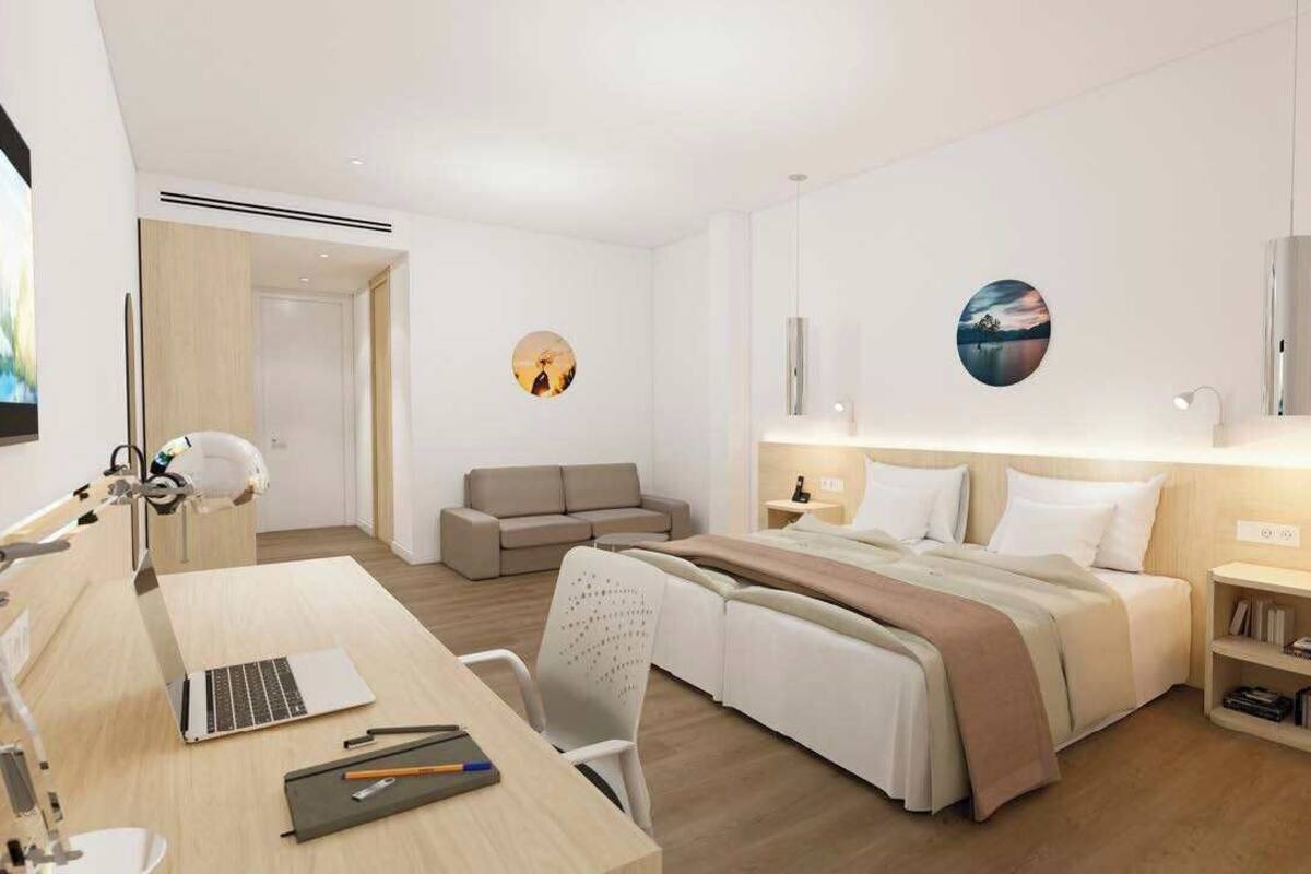 A modern hotel room with a bed, bedside tables, a desk with a laptop, a chair, and a sofa.