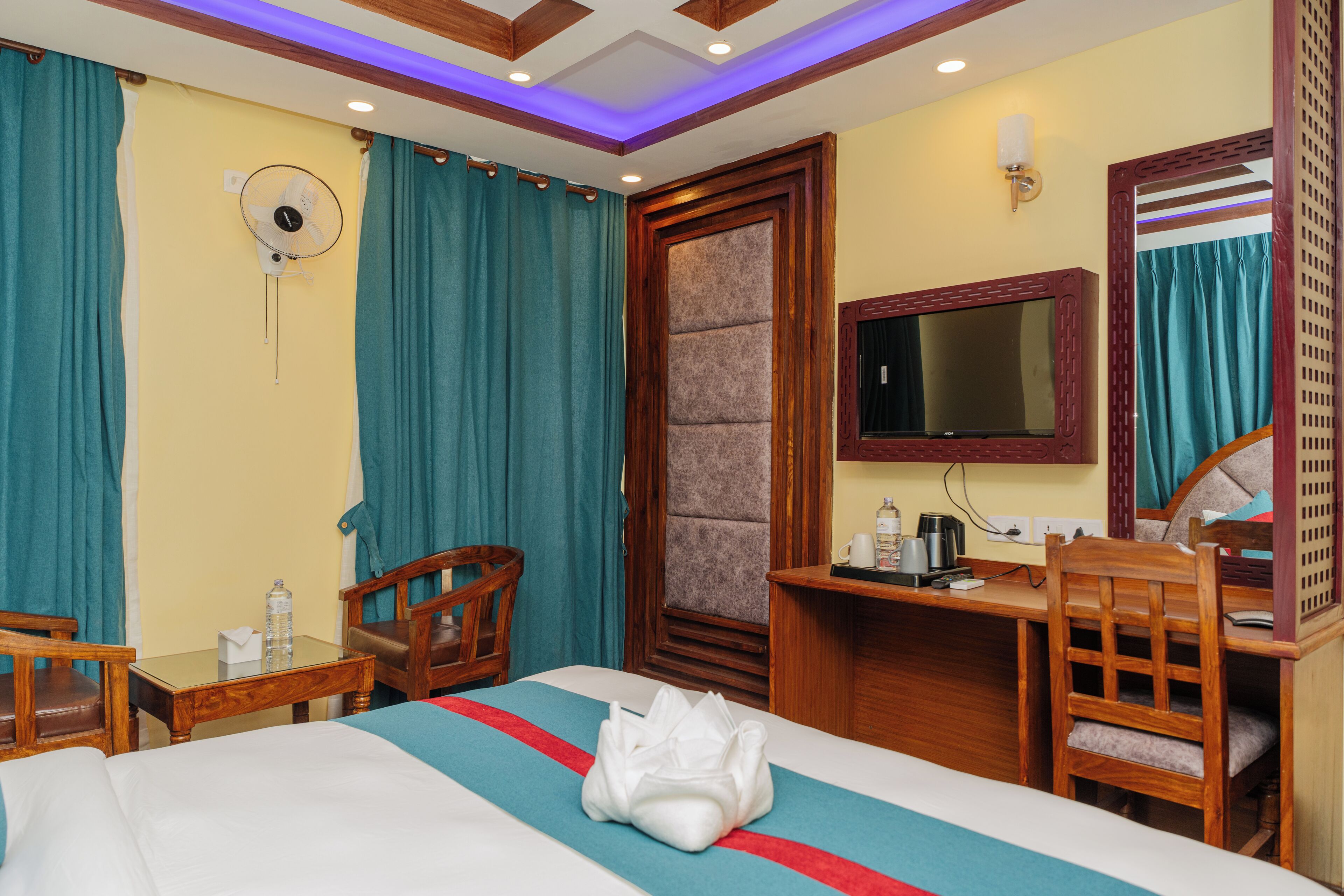 A hotel room with a bed, a desk, a chair, a TV, and a fan.
