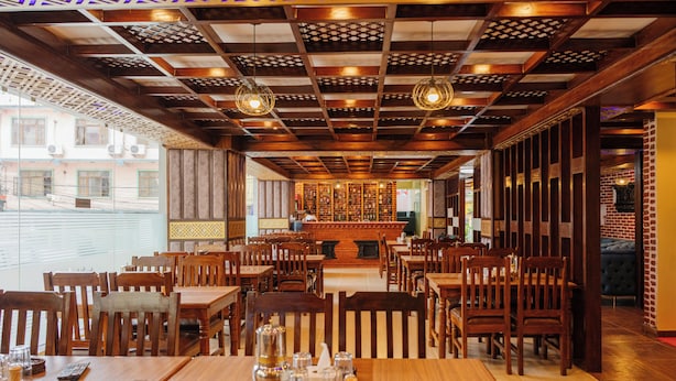 A restaurant with wooden tables and chairs, a patterned ceiling, and a brick wall. - Hotel Royal Airport (Kathmandu)