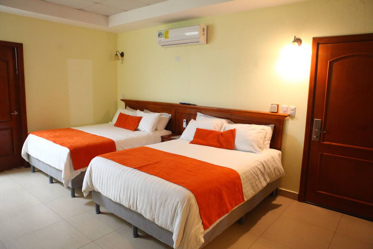 Une chambre d’hôtel avec deux lits, chacun doté de draps blancs et orange, d’une tête de lit en bois et d’un appareil de climatisation fixé au mur.