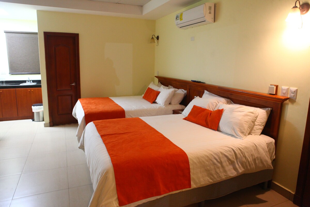 A hotel room with two beds, each with white bedding and orange accents, a wooden headboard, and a small bedside table.