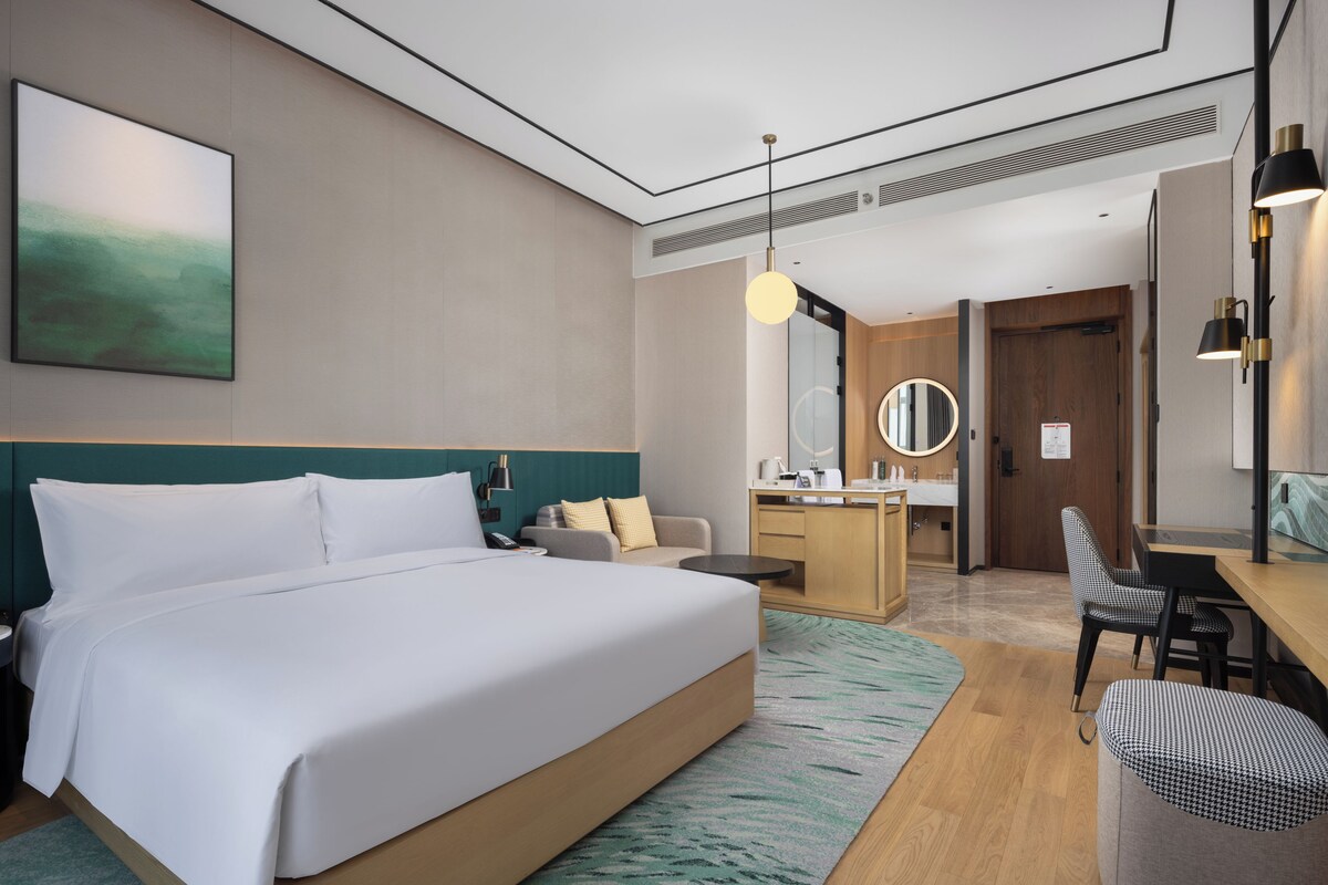 Ein modernes Hotelzimmer mit einem großen Bett, einem Schreibtisch, einem Sessel, einem runden Spiegel und einer kleinen Sitzecke.