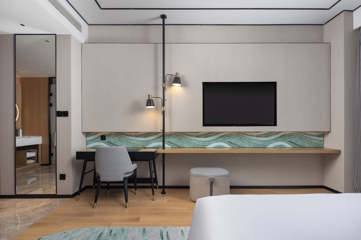 A modern hotel room with a bed, a desk with a chair, a TV mounted on the wall, and a patterned backsplash.
