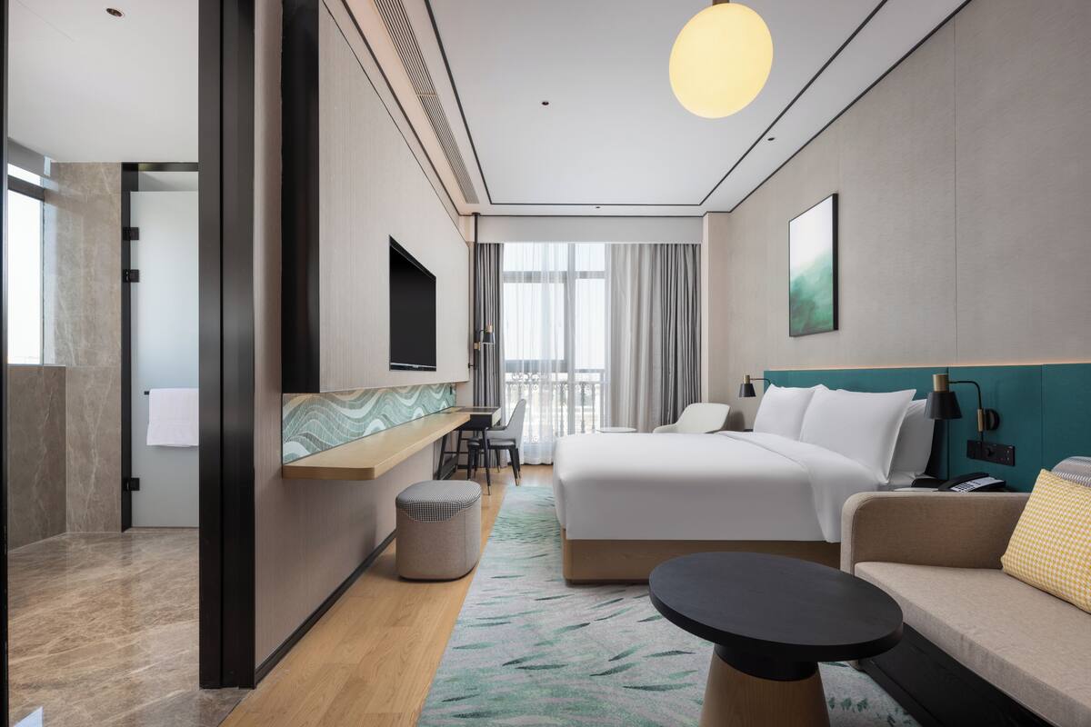 Una habitación de hotel moderna con una cama grande, un sofá, una mesita, un televisor y un balcón con cortinas.