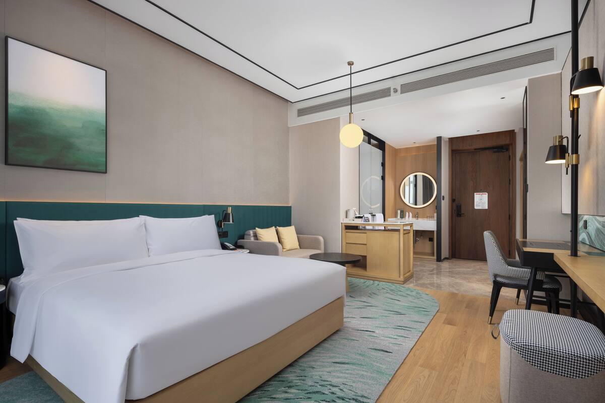 Una habitación de hotel moderna con una cama grande, un escritorio, una silla, un espejo redondo y una pequeña zona de estar.