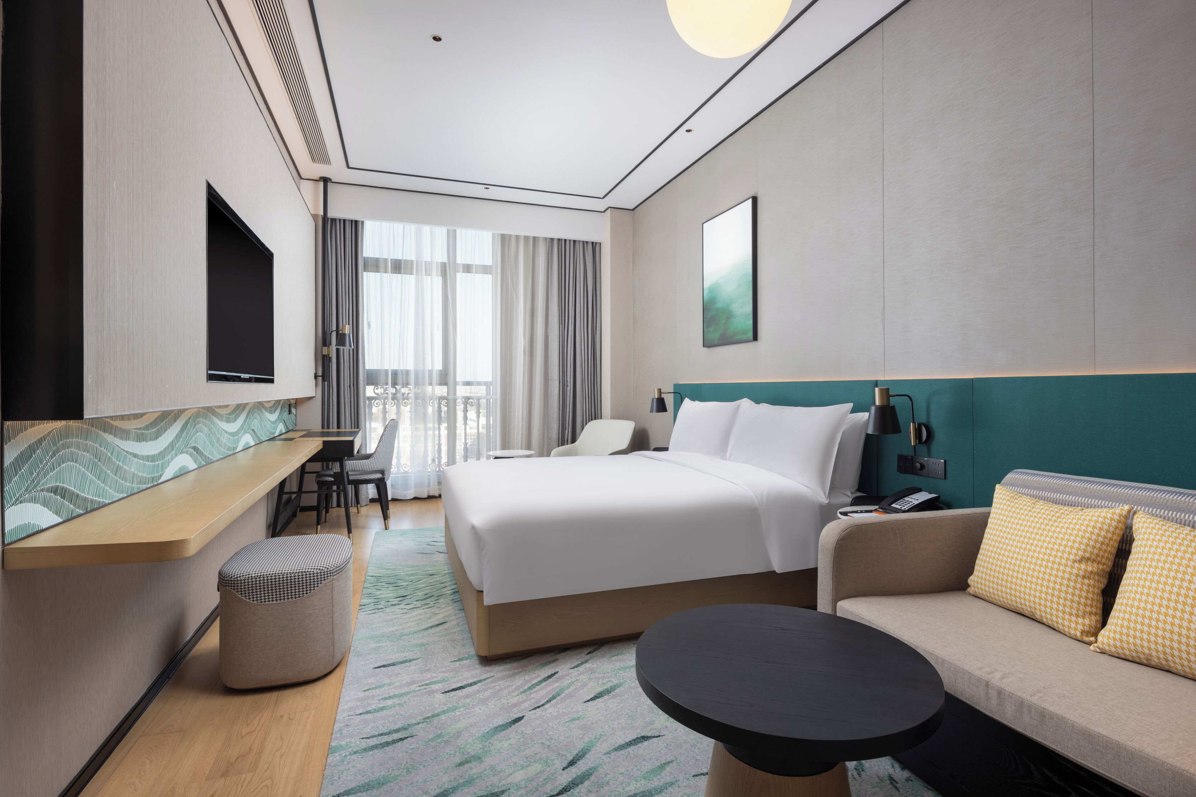 A modern hotel room with a large bed, a sofa, a small table, a TV, and a balcony with curtains.