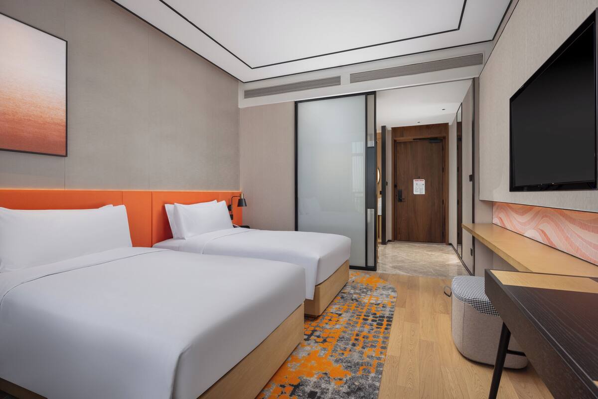 Habitación de hotel con dos camas, un escritorio, un televisor y un baño visible a través de una puerta de cristal.