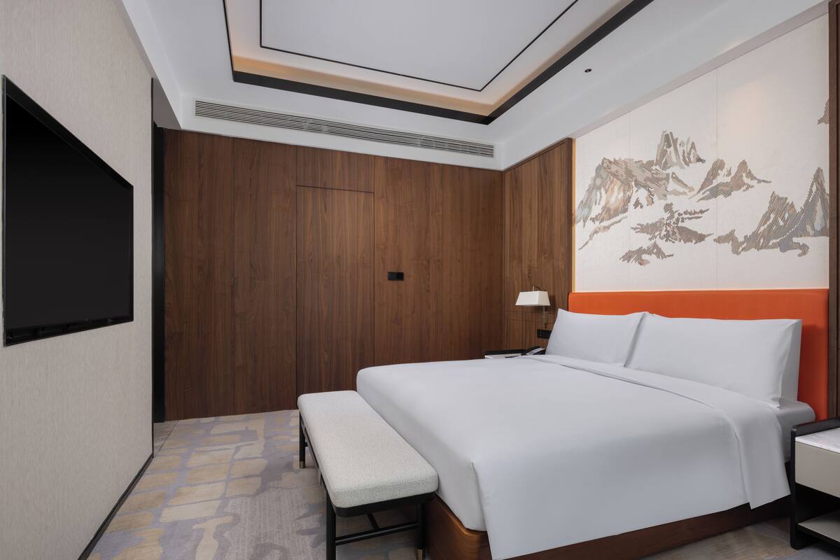Una habitación de hotel con una cama grande, un banco, un televisor de pantalla plana, una lámpara de pared y un adorno en la pared.