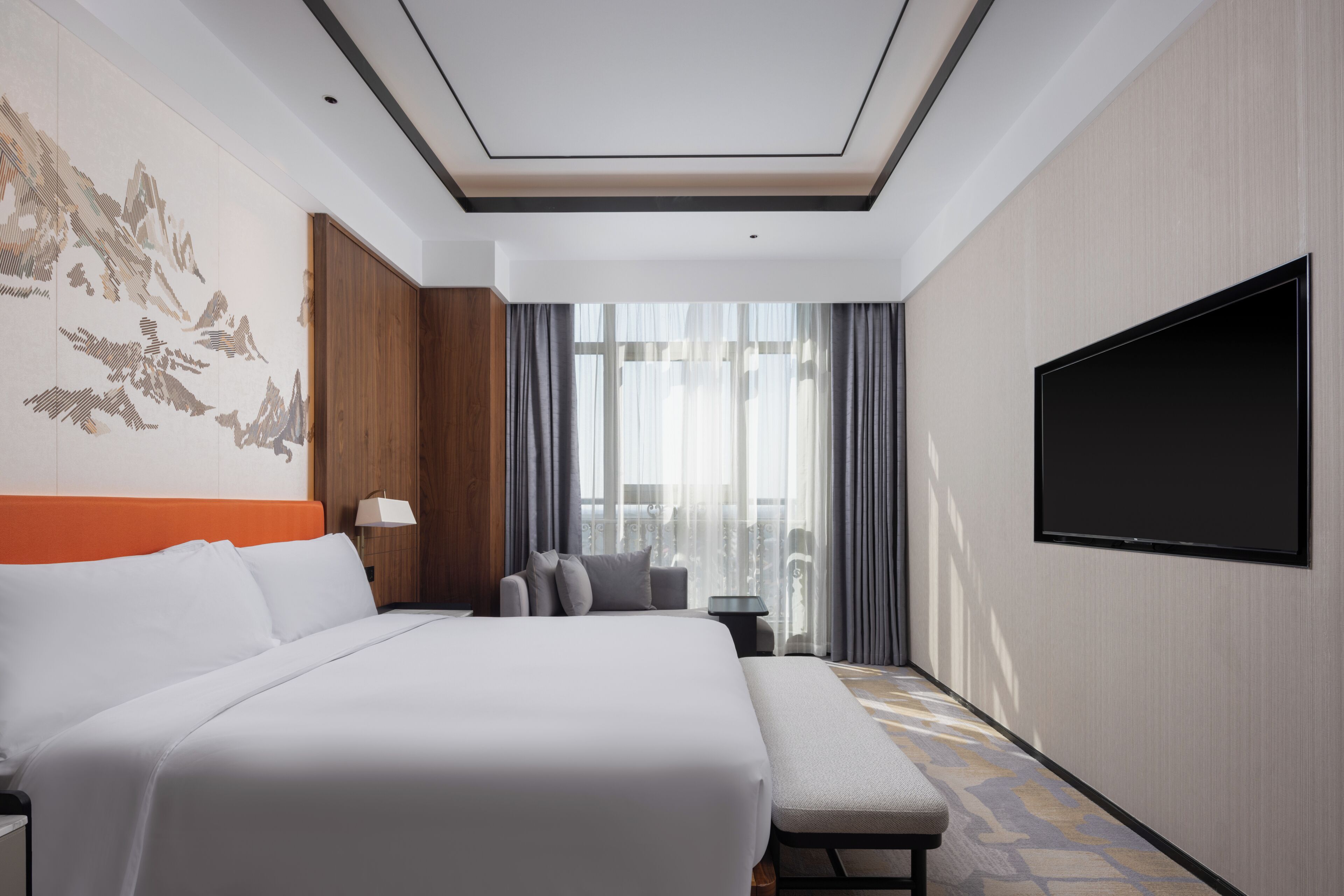 A hotel room with a large bed, a flat-screen TV, a bench, and a view of the city through large windows.